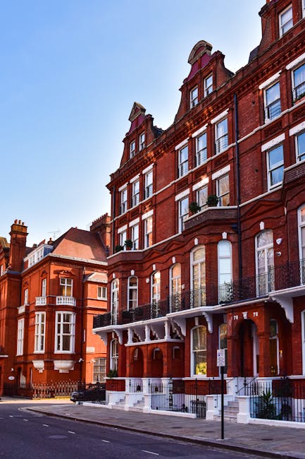 A row of historic red brick residential buildings with large sash windows and decorative white trim along Shepherd's Bush Road. The buildings feature balconies with black wrought iron railings and small potted plants. The street view includes a paved sidewalk, a lamppost, and a clear blue sky overhead. The exterior surfaces, particularly the brickwork and window frames, appear clean and well-maintained, reflecting a tidy and hygienic appearance. This image illustrates the type of property that may benefit from professional deep cleaning services provided by Shepherd's Bush Cleaners for end of tenancy preparations or general maintenance, ensuring surfaces such as brick facades and window areas are thoroughly cleaned and preserved.